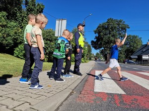 Zdjęcie przedstawia: chłopca przechodzącego przez pasy z ręką uniesioną do góry. Obok policjant i inne dzieci.