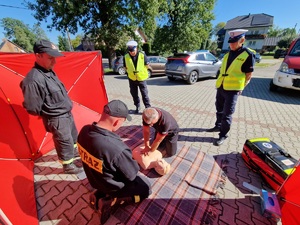 Zdjęcie przedstawia: mężczyznę podczas ćwiczeń przy fantomie, obok strażacy i policjanci.
