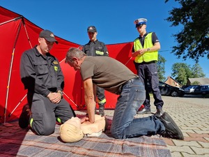 Zdjęcie przedstawia: mężczyznę podczas ćwiczeń udzielania pierwszej pomocy. Obok strażacy i policjant.