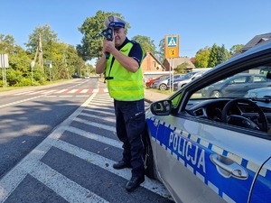 Zdjęcie przedstawia: umundurowanego policjanta podczas mierzenia prędkości samochodów, obok widoczny radiowóz.