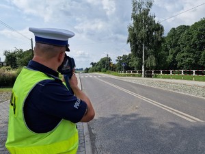 Zdjęcie przedstawia: policjanta podczas pomiaru prędkości nadjeżdżającego samochodu.