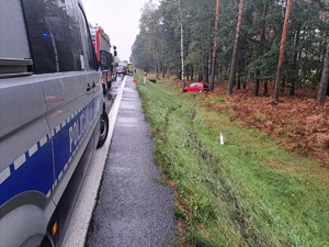 Zdjęcie przedstawia policyjny radiowóz i wozy straży pożarnej na jezdni. W przydrożnym rowie mały samochód osobowy.