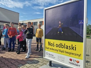 Zdjęcie przedstawia: duży plakat informujący o potrzebie noszenia odblasków. Z tyłu widoczna grupa dzieci.