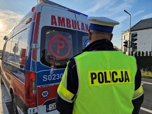 Zdjęcie przedstawia: policjanta stojącego przed karetką pogotowia.