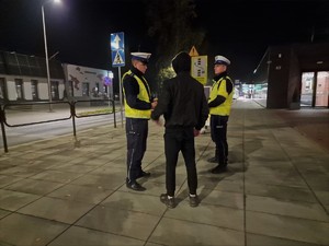 Zdjęcie przedstawia: umundurowanych policjantów podczas rozmowy z osobą ubraną całą na czarno.