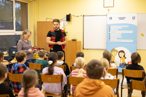 Zdjęcie przedstawia: ratownika medycznego podczas pogadanki o pierwszej pomocy. Przed mężczyzną dzieci siedzą na krzesełkach.