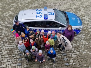 Zdjęcie przedstawia: grupę dzieci z opiekunami i policjantem. Z tyłu widoczny radiowóz policyjny.