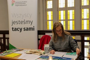 Zdjęcie przedstawia: kobietę przy stanowisku informacyjnym.