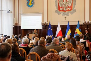 Zdjęcie przedstawia: kobietę na mównicy podczas przemówienia. Na sali siedzi mnóstwo osób.