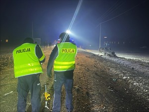 Zdjęcie. Dwóch umundurowanych policjantów podczas wykonywa czynności na miejscu wypadku drogowego. Za nimi uszkodzone samochody.