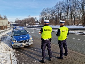 Zdjęcie przedstawia: policjantów ruchu drogowego. Za nimi widoczny oznakowany policyjny radiowóz.