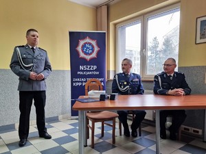 Zdjęcie przedstawia: trzech mundurowych. Jeden z policjantów stoi podczas przemówienia, dwóch siedzi przy stole.