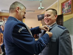 Zdjęcie przedstawia: uśmiechniętego policjanta, który otrzymuje wyróżnienie medalem od mundurowego.