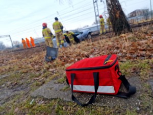 Zdjęcie przedstawia: z przodu apteczka, z tyłu uszkodzony samochód po zdarzeniu drogowym i strażacy.