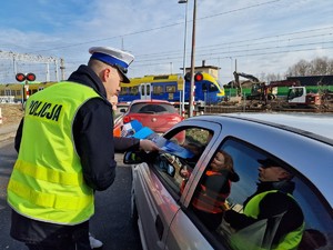 Zdjęcie przedstawia: umundurowanego policjanta i kobietę w kamizelce odblaskowej podczas rozmowy z kierowcą na zamkniętym przejeździe kolejowym. Z tyłu widoczny pociąg.