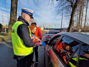 Zdjęcie przedstawia: uśmiechniętego policjanta, który stoi przy samochodzie osobowym. Widoczne kobiety w kamizelkach odblaskowych z gadżetami. Z tyłu widoczny pociąg.