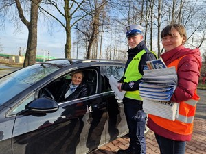 Zdjęcie przedstawia: kobietę za kierownicą samochodu, policjanta i kobietę w kamizelce odblaskowej z ulotkami, gazetkami i gadżetami.