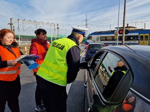 Zdjęcie przedstawia: policjanta i kobiety w kamizelkach odblaskowych podczas działań profilaktycznych na przejeździe kolejowym.