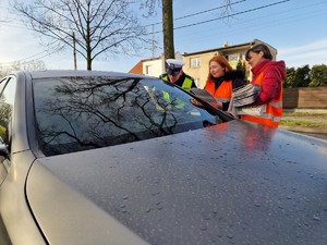 Zdjęcie przedstawia: samochód osobowy, umundurowanego policjanta i dwie kobiety w kamizelkach odblaskowych podczas rozmowy z kierowcą samochodu.