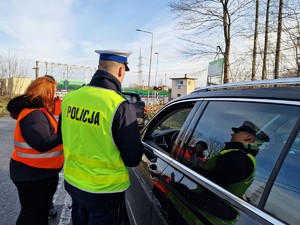 Zdjęcie przedstawia: policjanta i kobiety w kamizelkach odblaskowych podczas działań profilaktycznych. Z tyłu widoczny sygnalizator kolejowy.