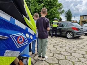 Zdjęcie przedstawia: znaczek policyjny, dalej dzieci i policjant podczas kontroli drogowej.