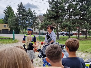 Zdjęcie przedstawia: mężczyznę który jest częstowany jabłuszkiem przez dziewczynkę. Wokół stoją dzieci.