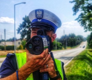 Zdjęcie przedstawia policjanta podczas pomiaru prędkości samochodów.