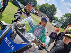 Zdjęcie przedstawia: dziewczynkę, która siedzi na policyjnym motocyklu, a obok stoi policjant. Mundurowy ma na głowie czapkę dziewczynki a ona jego.