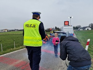 Zdjęcie przedstawia zdjęcie z planu. Widoczna mała dziewczynka do której podchodzi policjant.