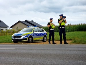 Zdjęcie przedstawia: dwóch policjantów ruchu drogoego stoi przy drodze, z tyłu widoczny radiowóz.