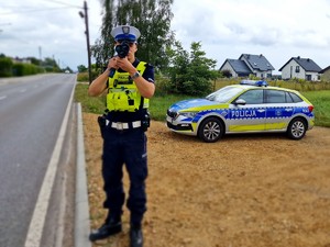 Zdjęcie przedstawia: policjanta podczas pomiaru prędkości, z tyłu widoczny radiowóz.