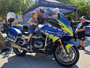 Zdjęcie przedstawia policjanta, który sadza na policyjny motocykl małego chłopca.