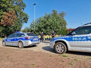 Zdjęcie przedstawia: dwa policyjne radiowozy, z tyłu widoczni policjanci i dzieci.