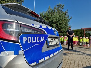 Zdjęcie przedstawia: tylną część policyjnego radiowozu, za nim widoczni policjant i dzieci.