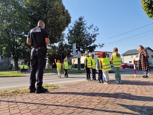 Zdjęcie przedstawia: policjanta z dziećmi podczas ćwiczenia przechodzenia przez przejście dla pieszych.