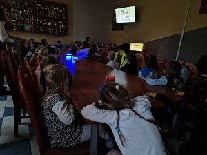 Zdjęcie przedstawia: 
Przedszkolaki siedzące przy stole oglądające bajkę edukacyjną na telewizorze.