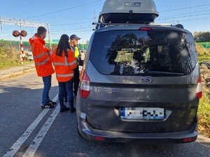 Zdjęcie przedstawia: 
Tył samochodu, stojącego przed przejazdem kolejowym, oraz umundurowanego policjanta wraz z przedstawicielami Kolei Śląskich.