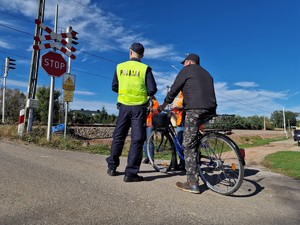 Zdjęcie przedstawia: 
Rowerzystę stojącego przed przejazdem kolejowym rozmawiającego z umundurowanym policjantem oraz dwóch przedstawicieli Kolei Śląskich.