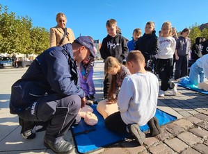 Na zdjęciu umundurowany policjant wraz z dziećmi ćwiczącymi prowadzenie resuscytację na fantomie