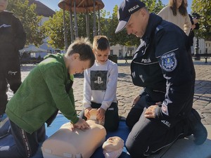 Na zdjęciu umundurowany policjant wraz z dziećmi ćwiczącymi prowadzenie resuscytację na fantomie