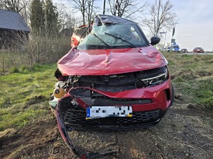 Zdjęcie przedstawia: czerwony pojazd osobowy widziany od przodu roztrzaskany z swojej prawej strony
