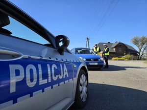 Zdjęcie przedstawia: radiowóz a w tle dwóch funkcjonariuszy policji rozmawiających z kierowcą niebieskiej osobówki