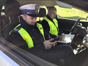 Zdjęcie przedstawia: dwóch funkcjonariuszy policji w radiowozie, spisujących notatkę