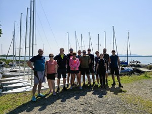 Zdjęcie przedstawia: grupę ludzi strojach sportowych stojących w porcie obok żaglówek.