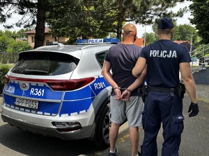 Zdjęcie przedstawia: mężczyznę, który ma na rękach kajdanki i jest trzymany przez policjanta.