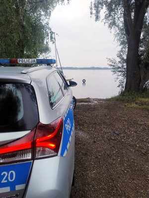 Zdjęcie przedstawia: auto policyjne stojące przy linii brzegu zbiornika wodnego.