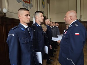 Zdjęcie przedstawia: policjanta stojącego na przeciwko szeregu policjantów.