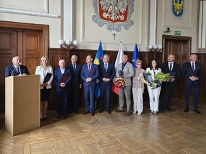 Zdjęcie przedstawia: grupę ludzi stojących obok osoby, która przemawia na mównicy.