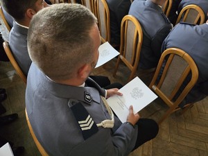 Zdjęcie przedstawia: siedzącego policjanta z teczką w ręce.