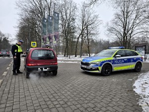Zdjęcie przedstawia umundurowanego policjanta, oznakowany radiowóz oraz samochód osobowy.